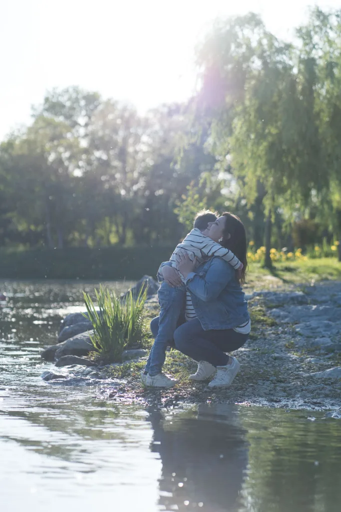 Une personne s'agenouille au bord de l'eau, serre dans ses bras un enfant entouré de verdure et de soleil, capturant un moment magnifique, parfait pour la photographie de style de vie dans les Landes pittoresques.