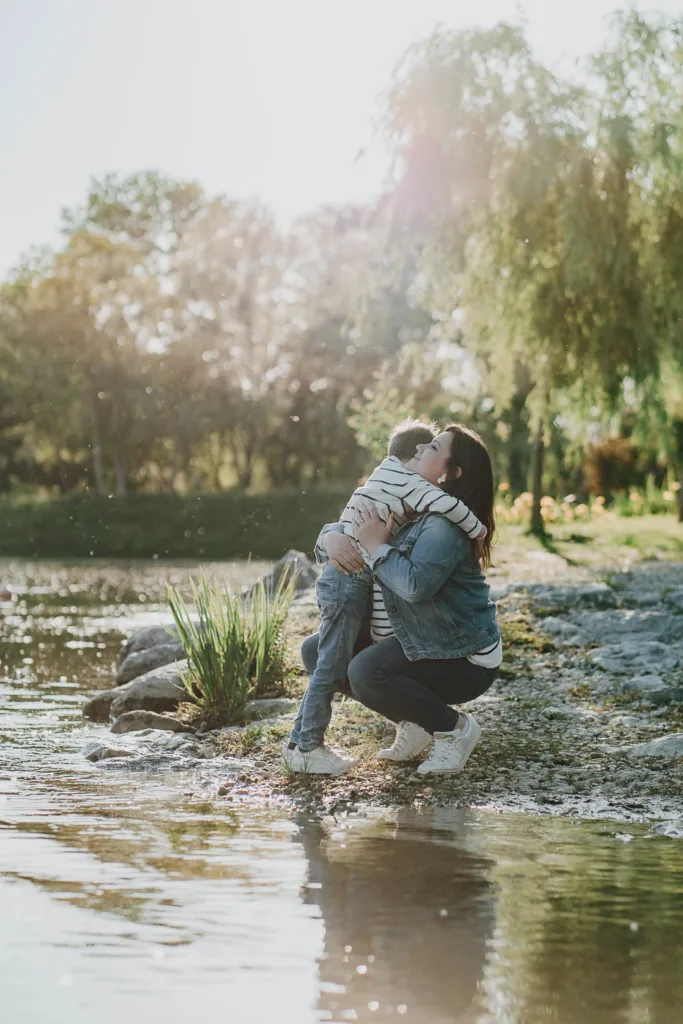 Un adulte et un enfant partagent une étreinte près d’un plan d’eau entouré d’arbres et de verdure par une journée ensoleillée, capturant un moment parfait de leur style de vie.