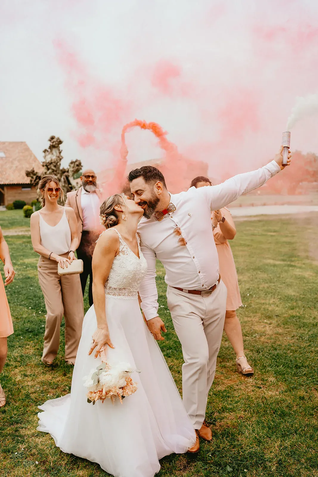 Des mariés s'embrassent sur un terrain herbeux avec des amis autour d'eux tandis que des fusées éclairantes de fumée magenta sont en arrière-plan. La mariée tient un bouquet et le marié lève un bras tenant une fusée éclairante. Cette scène est une pure inspiration de mariage pour tout couple planifiant son grand jour.