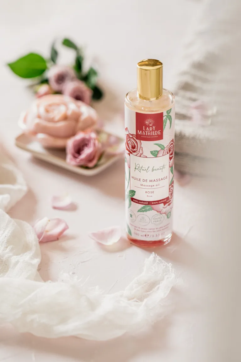 Un flacon d'huile de massage Lady Mathilde à l'extrait de rose se tient debout sur une surface légère. Autour de lui se trouvent des pétales de rose, une fleur de rose et un tissu texturé, le tout magnifiquement capturé par un photographe professionnel landais.