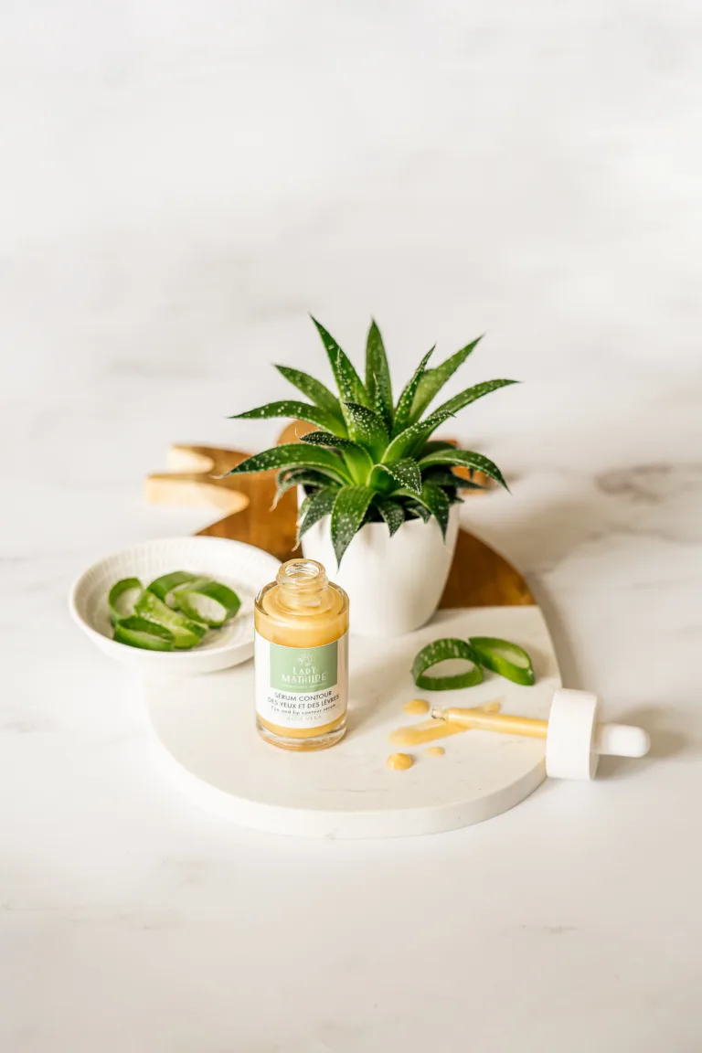 Un flacon de sérum de soin est ouvert sur un plateau rond blanc avec son compte-gouttes à proximité. Capturée par un photographe professionnel landais, la scène est complétée par une petite plante succulente en pot et un bol avec des feuilles d'aloès tranchées en arrière-plan.