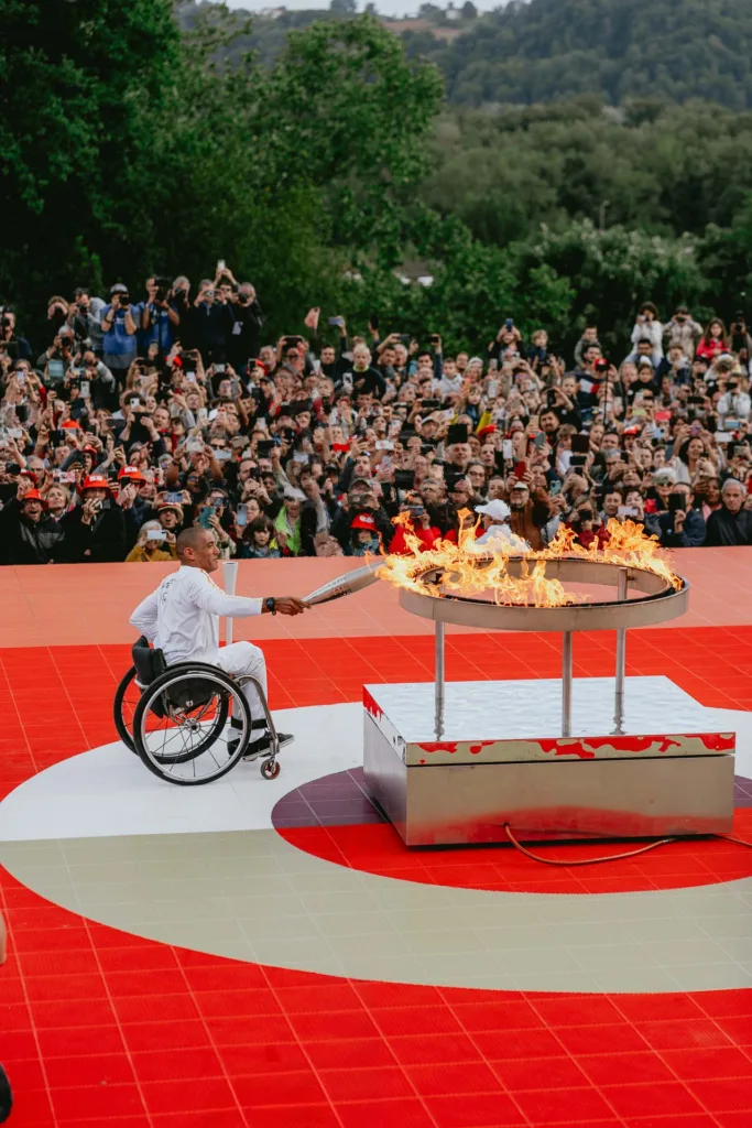 Une personne en fauteuil roulant allume une flamme de cérémonie avec une torche, entourée d'une foule de badauds, dont un photographe professionnel landais capturant l'instant.
