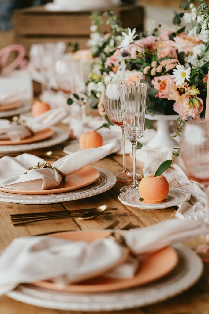 Table élégante avec assiettes roses, serviettes blanches, couverts dorés, verres en cristal et centres de table fleuris. S'inspirant d'un thème de mariage, une pêche magenta est placée sur chaque assiette.