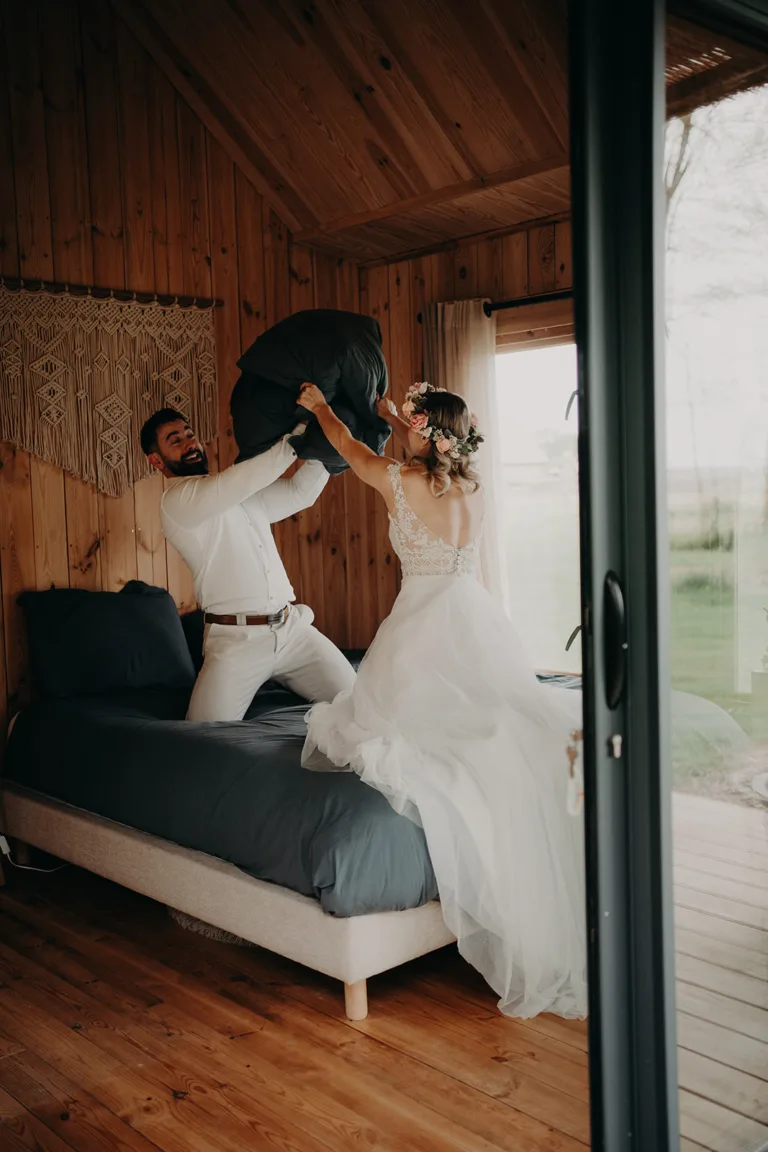 Une mariée vêtue d'une robe de mariée blanche s'engage de manière ludique dans une bataille d'oreillers avec un homme vêtu d'une chemise et d'un pantalon blancs, à l'intérieur d'une cabane en bois avec une grande fenêtre. La scène chaleureuse capture le moment joyeux du couple, entouré par la beauté sereine des Pyrénées à l'extérieur.