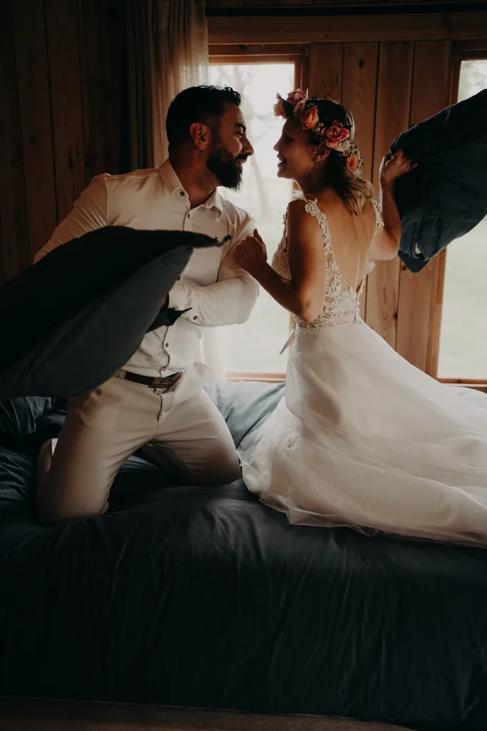 Un couple en tenue de mariage se livre de manière ludique à une bataille d'oreillers sur un lit, avec des murs en bois et une fenêtre donnant sur les Pyrénées en arrière-plan.