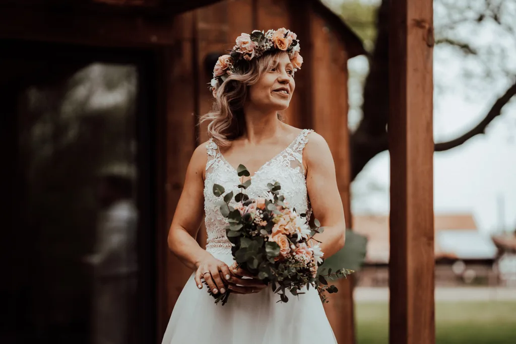Une mariée portant une couronne florale et tenant un bouquet se tient à l'extérieur près d'une structure rustique en bois dans les Pyrénées. Elle sourit et regarde à sa gauche.