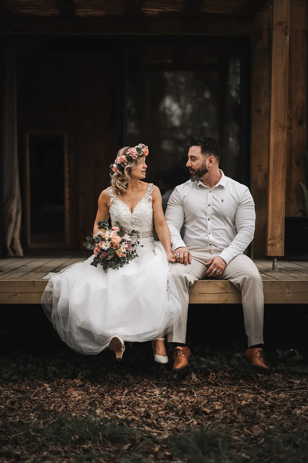 Une mariée vêtue d'une robe blanche et d'un casque fleuri est assise à côté d'un marié vêtu d'une chemise blanche et d'un pantalon kaki sur une plate-forme en bois, se tenant la main et se regardant avec des expressions calmes, leur moment serein rappelant une vieille photo de séance.