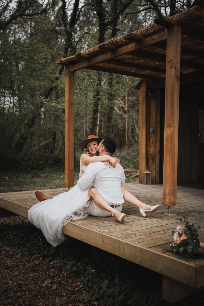 Un couple vêtu d'une tenue de mariage blanche est assis au bord d'une terrasse en bois dans une zone boisée des Pyrénées, souriant et s'embrassant. Un bouquet repose par terre à proximité.