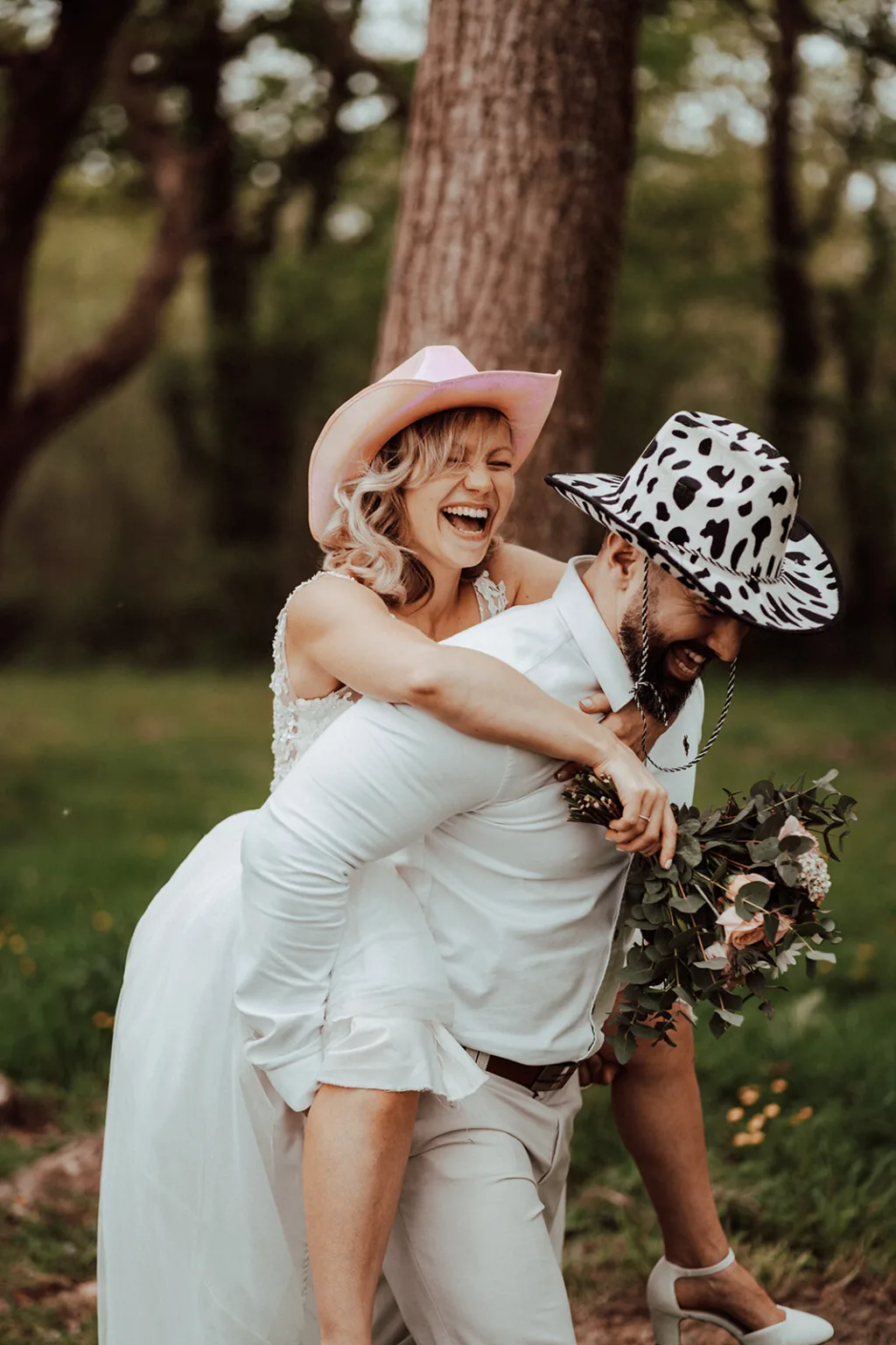 Une mariée, portant un chapeau rose, chevauche joyeusement son marié, qui arbore un chapeau à imprimé vache. Sur cette photo fantaisiste prise dans la région boisée des Pyrénées, la mariée tient un bouquet de fleurs tandis que leurs rires résonnent à travers les arbres.