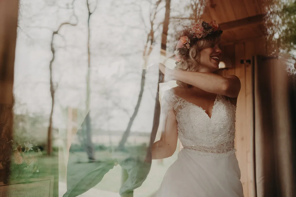 Une mariée vêtue d'une robe blanche et d'un bandeau fleuri se tient près d'une fenêtre dans les Pyrénées, souriante et levant le bras.