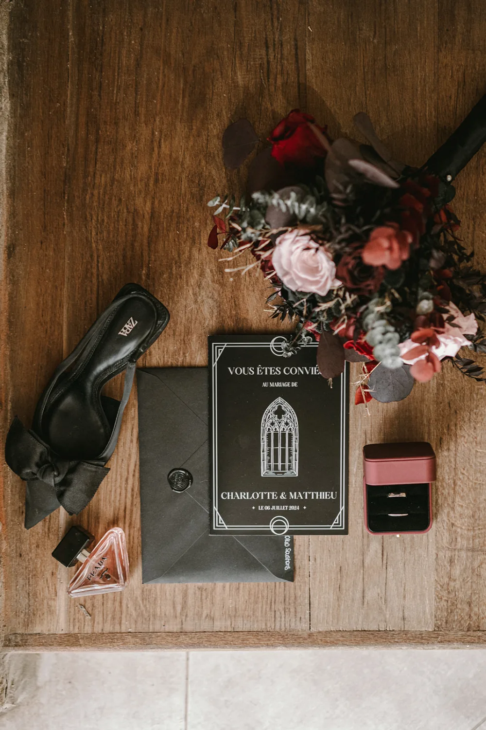 Un superbe bouquet floral, une chaussure à talons noirs, une carte d'invitation élégante, une boîte à bijoux contenant des alliances et un flacon de parfum sont soigneusement disposés sur une surface en bois—offrant ainsi une scène idéale pour un photographe de mariage ou famille dans les Landes.