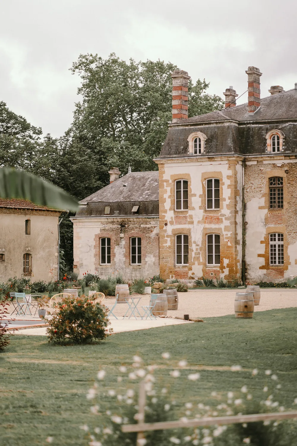 Une vaste bâtisse historique aux hautes cheminées, entourée d'arbres et d'un jardin bien entretenu, avec en scène plusieurs chaises et tables colorées au premier plan, formant un cadre idéal pour un photographe de mariage ou de famille dans la région charmante des Landes. .