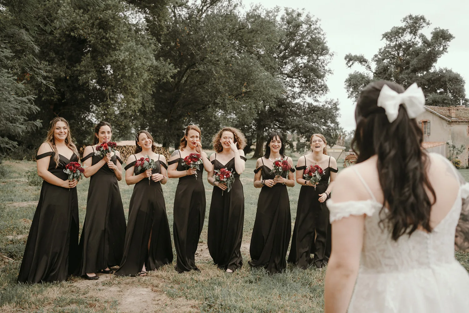 Sur une pelouse verdoyante bordée d'arbres, une mariée vue de dos fait face à ses huit demoiselles d'honneur vêtues de robes noires et tenant des bouquets rouges, un instant parfait capturé par un photographe de mariage et famille dans les Landes.