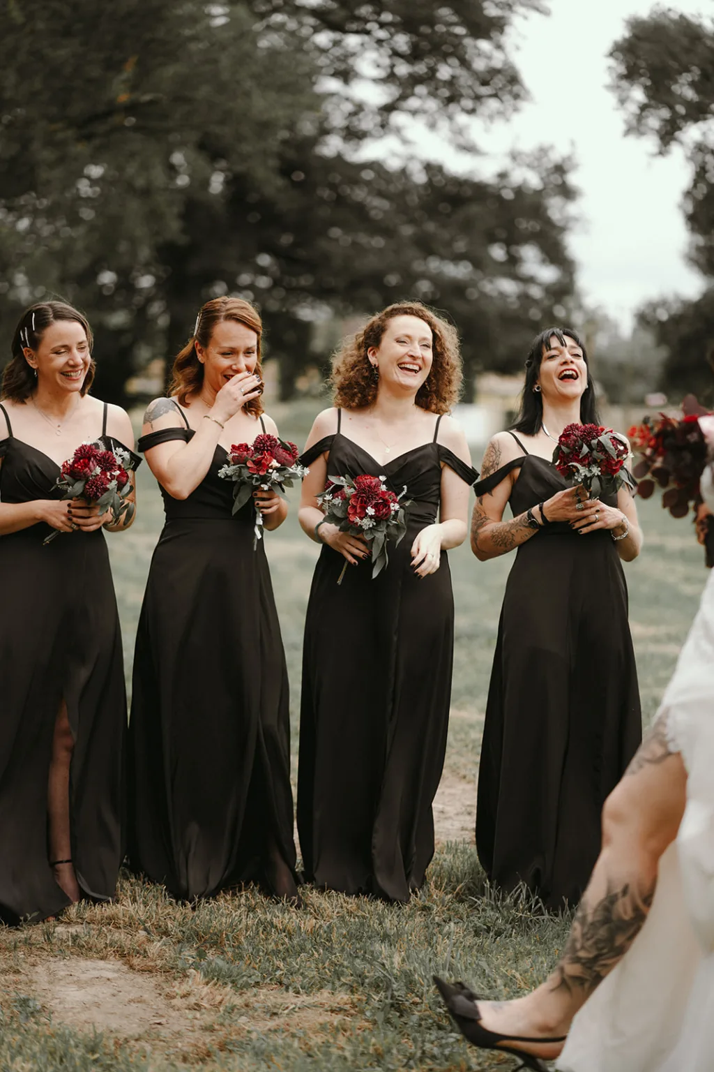 Sous un ciel dégagé et entourées par une nature luxuriante, quatre femmes habillées de robes noires assorties tiennent des bouquets de fleurs rouges éclatantes tout en riant avec bonheur, une scène chaleureusement immortalisée par un photographe de famille dans les Landes spécialisé dans les photos en plein air , capturant la beauté naturelle des arbres et des étendues d'herbe environnante.