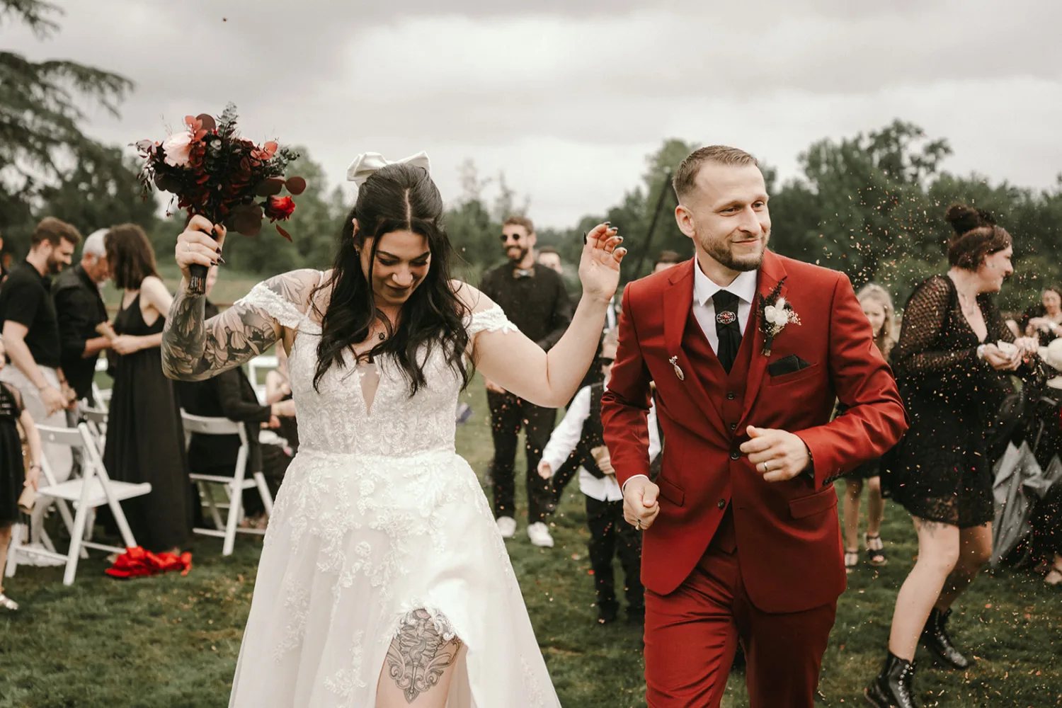 Un couple heureux descend une allée extérieure, la mariée vêtue d'une robe blanche tenant un bouquet et le marié en costume rouge, tandis que les invités applaudissent et jettent des confettis en arrière-plan, capturant ainsi des moments parfaits pour tout photographe mariage ou photographe famille dans les Landes.