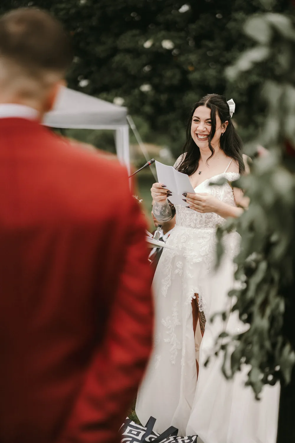 Une mariée en robe blanche de mariage allumée un discours tout en souriant, debout à l'extérieur à côté d'un marié vêtu d'une veste de costume rouge, dans un cadre idyllique et naturel magnifiquement immortalisé par un photographe de famille des Landes spécialisé en photographie de mariage.