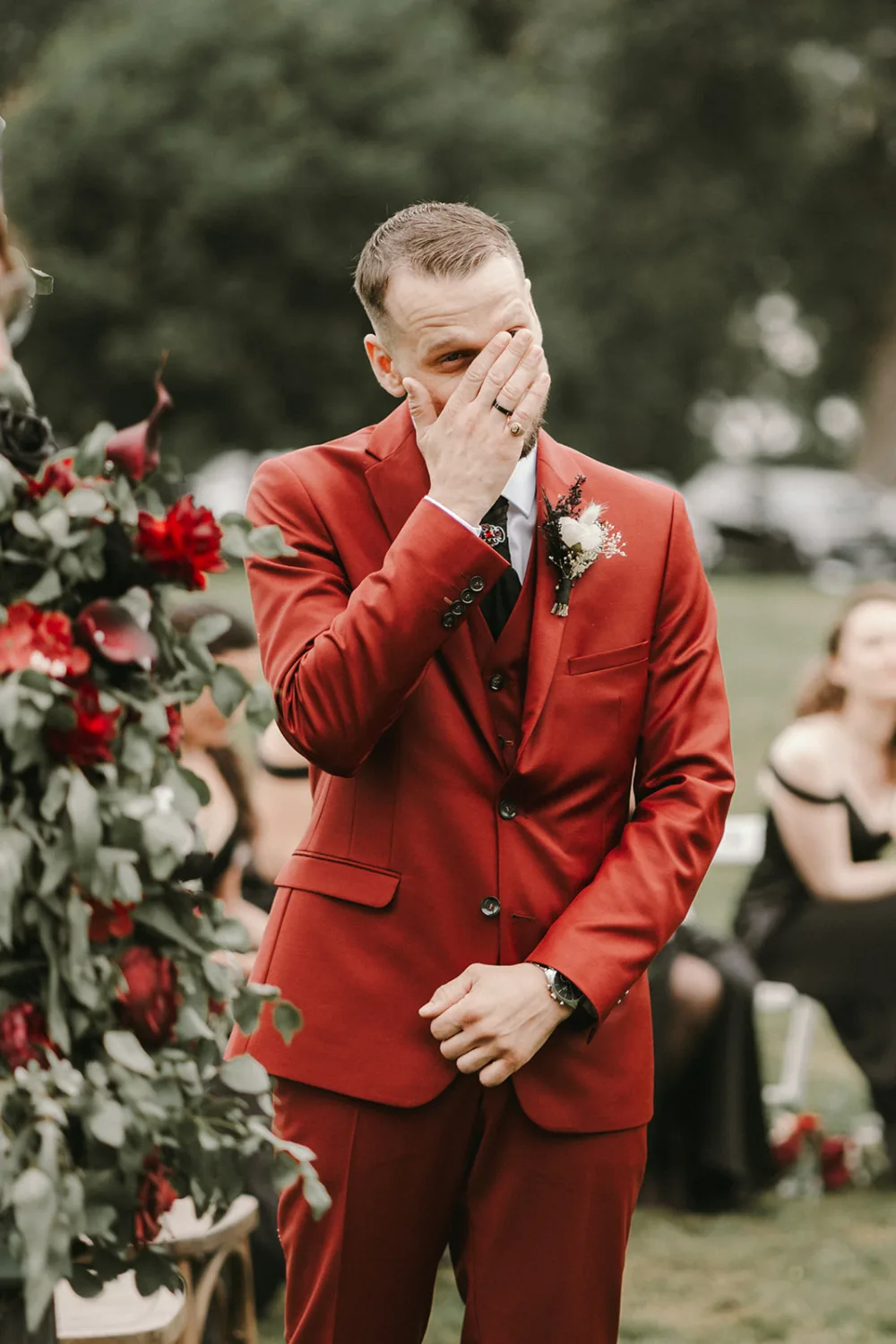 Un individu en costume rouge, comprenant son visage d'une main, se tient à l'extérieur près d'une magnifique composition florale, entouré de convives assis en arrière-plan, créant une scène idéale pour un photographe de mariage ou de famille dans les Landais.