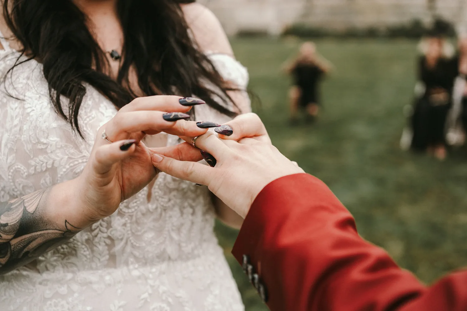 Dans cette scène capturée par un talentueux photographe de mariage ou de famille des Landes, une personne en robe blanche en dentelle place délicatement une bague au doigt d'une autre personne vêtue d'une veste rouge, le tout dans un cadre enchanteur en extérieur avec des silhouettes floues en arrière-plan ajoutant à l'atmosphère romantique et authentique propre aux célébrations familiales dans les magnifiques paysages des Landes.