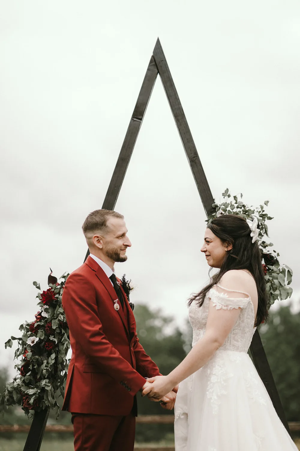 Lors de leur cérémonie de mariage, un couple se tient la main devant une arche triangulaire en bois ornée de fleurs rouges et blanches, le marié portant un costume rouge et la mariée une robe blanche, capturée avec amour par un photographe de famille dans les Landes. .