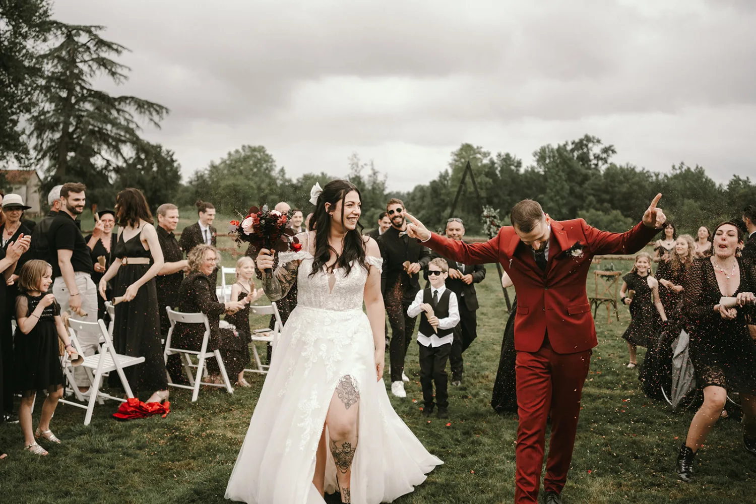 Un couple de mariés, la mariée en robe blanche et le marié en costume rouge, célèbrent leur union en descendant une allée herbeuse entourée d'invités applaudissant et acclamant sous un magnifique décor boisé—un instant parfait immortalisé par le photographe de mariage et famille des Landais.