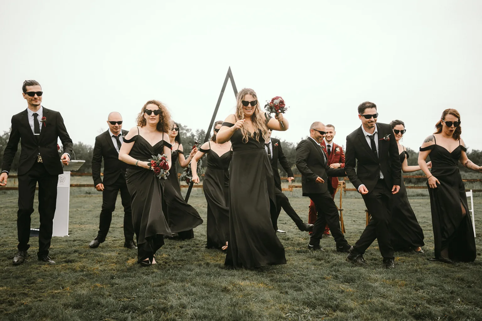 Un groupe de personnes vêtues d'élégants habits noirs et portant des lunettes de soleil se tient en plein air sur une étendue herbeuse, évoquant le charme d'une séance de photographie familiale dans les Landes ; Certains membres du groupe observent des fleurs et présentent de manière variée, présentant l'atmosphère unique et chaleureuse que seul un photographe de mariage ou une famille en terres landaises peut capturer.