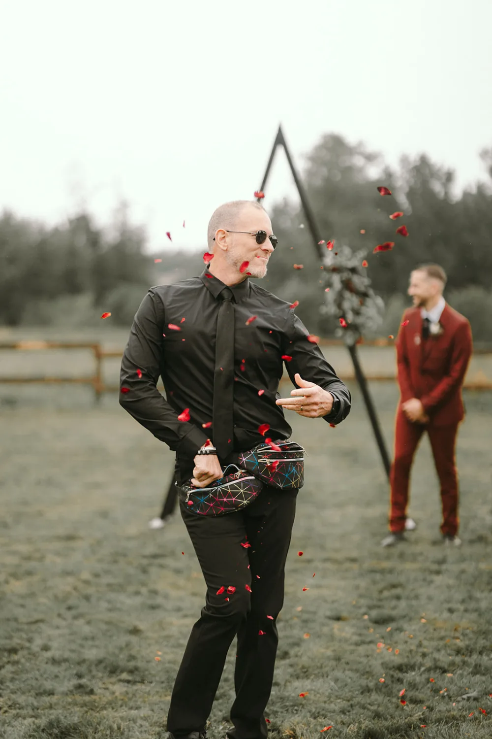 Lors d'un événement en plein air magnifiquement capturé par le photographe famille Landes, un homme vêtu de noir et portant des lunettes de soleil jet des pétales de fleurs depuis une banane, tandis qu'une autre personne en costume rouge se tient en arrière- plan, créant ainsi un moment de joie partagé idéal pour les photos de mariage dans les Landes.