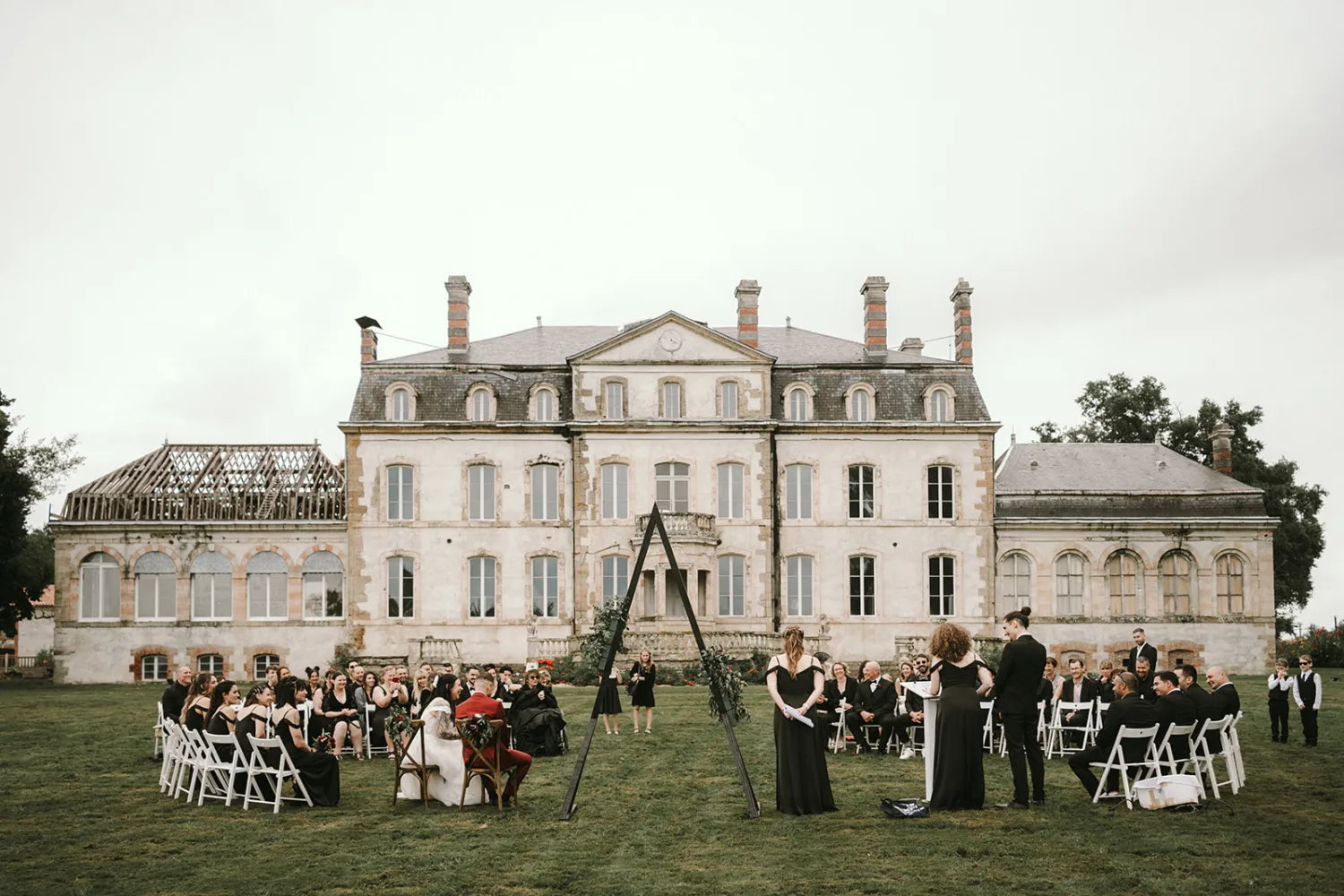 Cette cérémonie de mariage en plein air devant un grand manoir historique, avec des invités assis sur des chaises pliantes blanches et face à une arche en bois triangulaire, offre un cadre idyllique pour un photographe de mariage ou famille dans les Landes désireux de saisir des moments précieux et intemporels.