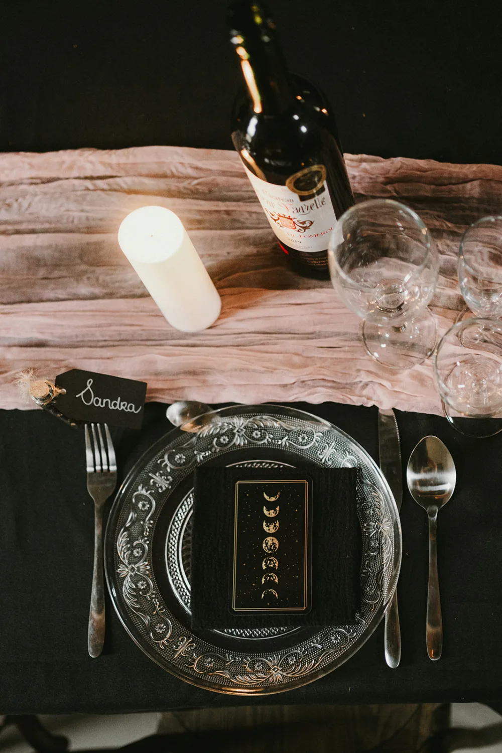 Cette image capturée par un photographe de mariage ou de famille dans les Landes présente une somptueuse mise en scène de table avec une bougie, une bouteille de vin, deux verres à vin vides et des couverts disposés sur une nappe noire; au centre du décor se trouve une serviette pliée accompagnée d'une carte illustrant les différentes phases de la lune, soigneusement placée sur une assiette en verre transparent, ajoutant une touche élégante et personnelle avec un prénom "Sandra" inscrit sur un petit tag nominatif.