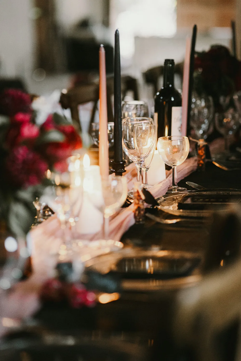 Une table à manger faiblement éclairée ornée de verres à vin, d'une bouteille de vin, de bougies et de centres de table floraux, est parfaitement dressée pour un rassemblement formel ou un événement, créant ainsi une ambiance élégante et accueillante que même un * photographe mariage Landes* ou *photographe famille Landes* trouverait enchanteur pour capturer des moments inoubliables.