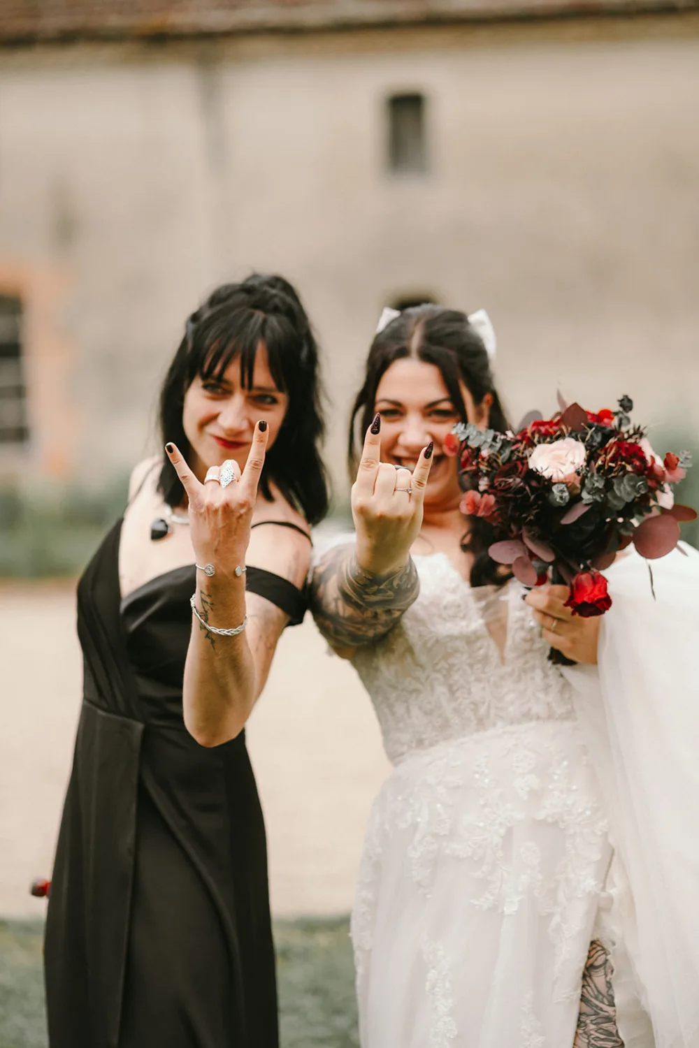 Dans cette image captivante prise par un photographe de mariage et famille dans les Landes, deux femmes présentent en extérieur : l'une arborant une magnifique robe de mariée blanche avec un bouquet à la main, l'autre vêtue d'une élégante robe sombre; toutes deux font le geste "rock on" des mains au milieu d'un paysage époustouflant qui souligne la beauté naturelle et romantique de la région.