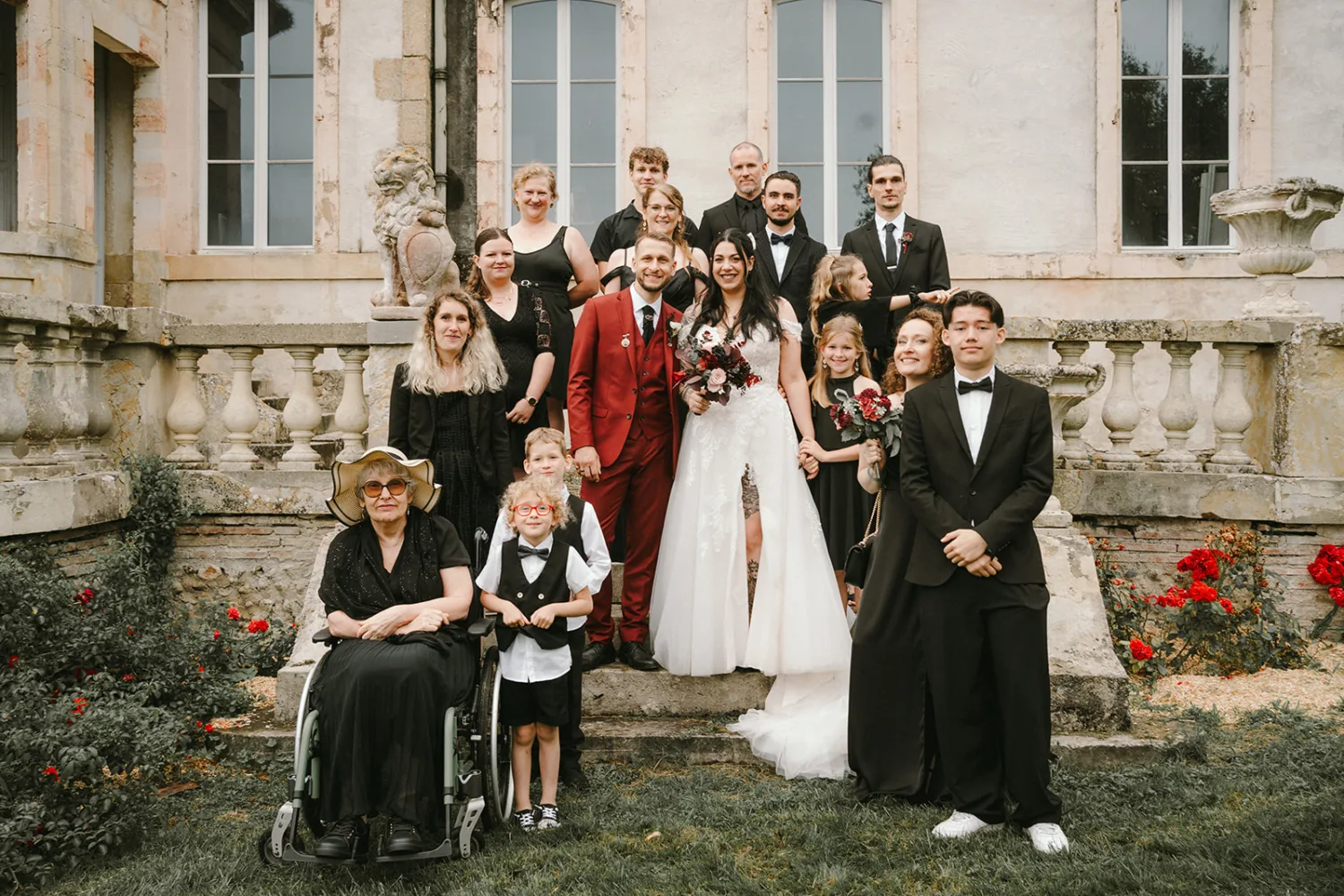 Dans cette magnifique photo de groupe de mariage capturée par un talentueux photographe famille Landes, le marié d'un élégant costume rouge et la mariée resplendissante dans sa robe blanche prennent place au centre, entourés de leurs proches et amis qui posent joyeusement sur les marches. d'un grand bâtiment.