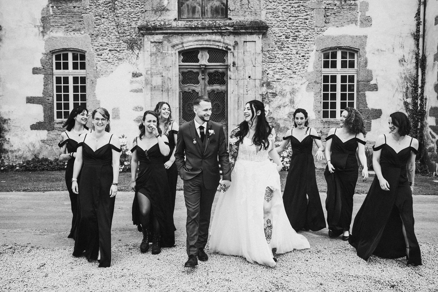 Une mariée et un marié marchent main dans la main, entourés de demoiselles d'honneur en robes noires, tandis que le *photographe de mariage et famille dans les Landes* immortalise cet instant devant un bâtiment en pierre.