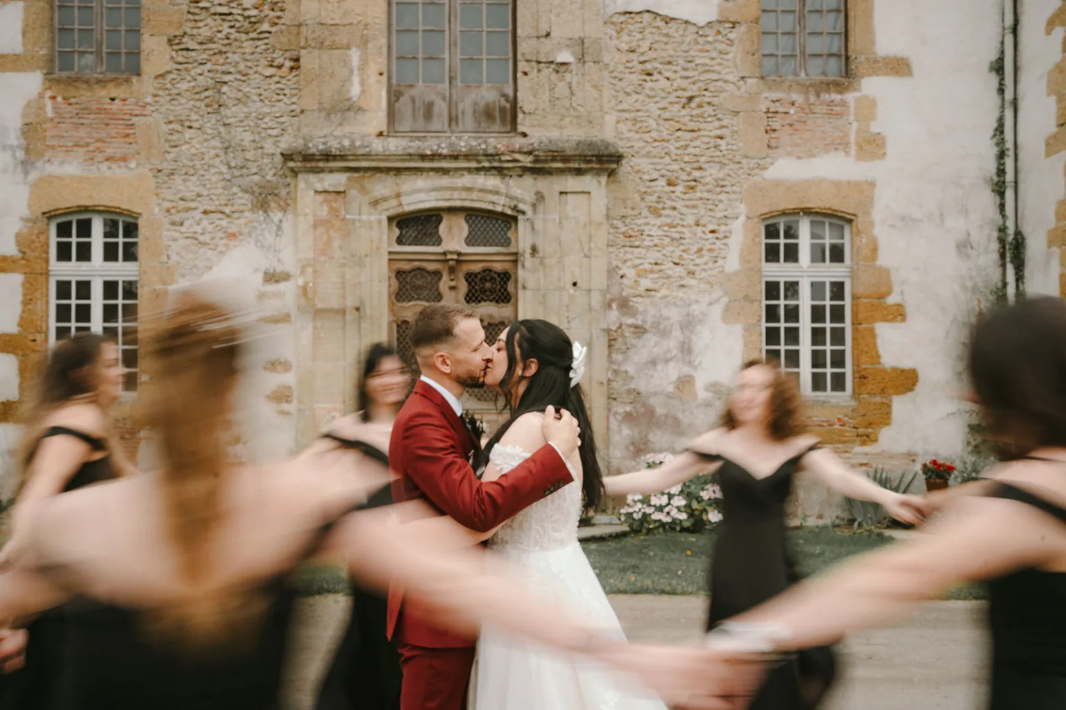 À travers l'objectif d'un photographe de mariage talentueux spécialisé en photographe de famille dans les Landes, cette image captivante saisit un moment magique où la mariée et le marié s'embrassent tendrement devant un vieux bâtiment pittoresque, tandis que des demoiselles d'honneur vêtues de robes noires forment une ronde autour d'eux, créant un effet de mouvement flou qui ajoute une touche artistique à cette scène inoubliable.