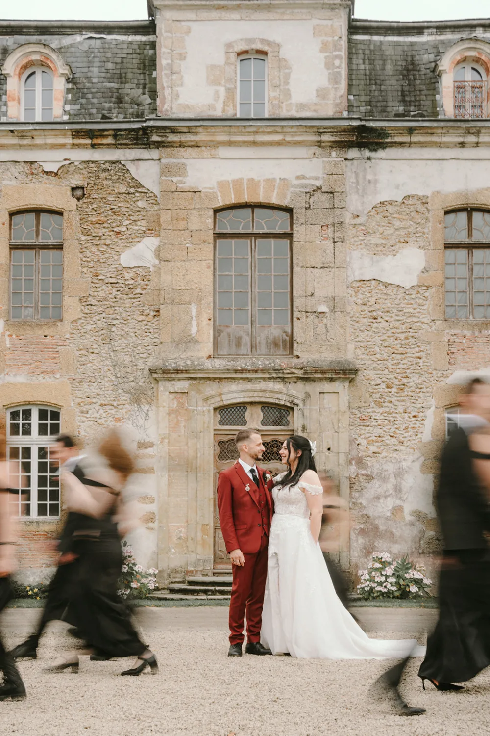 Un couple, vêtu d'un costume rouge et d'une robe de mariée blanche, se tient devant un vieux bâtiment en pierre alors que des silhouettes floues passent devant eux, immortalisant un moment intemporel digne de toute précieuse collection de photographies de mariage ou de famille dans les Landes par un photographe professionnel.