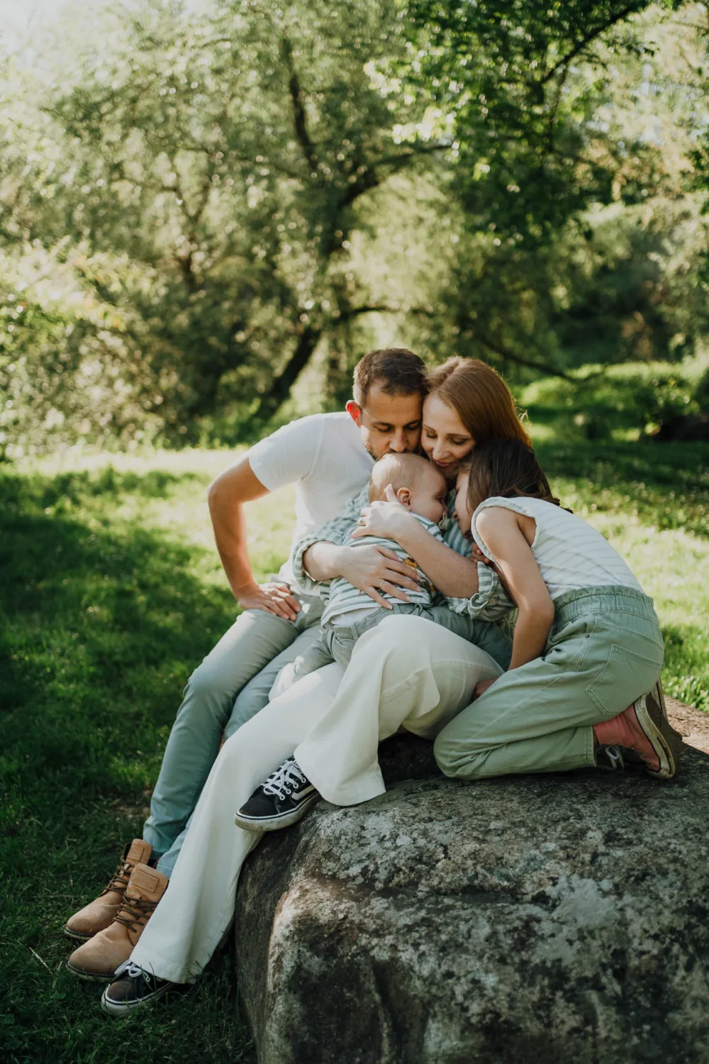 Une famille de trois personnes est assise étroitement ensemble sur un gros rocher dans un parc verdoyant et luxuriant. Les parents embrassent leur enfant, créant une scène chaleureuse et intime qui pourrait être la capture parfaite pour toute famille photographe des Landes.