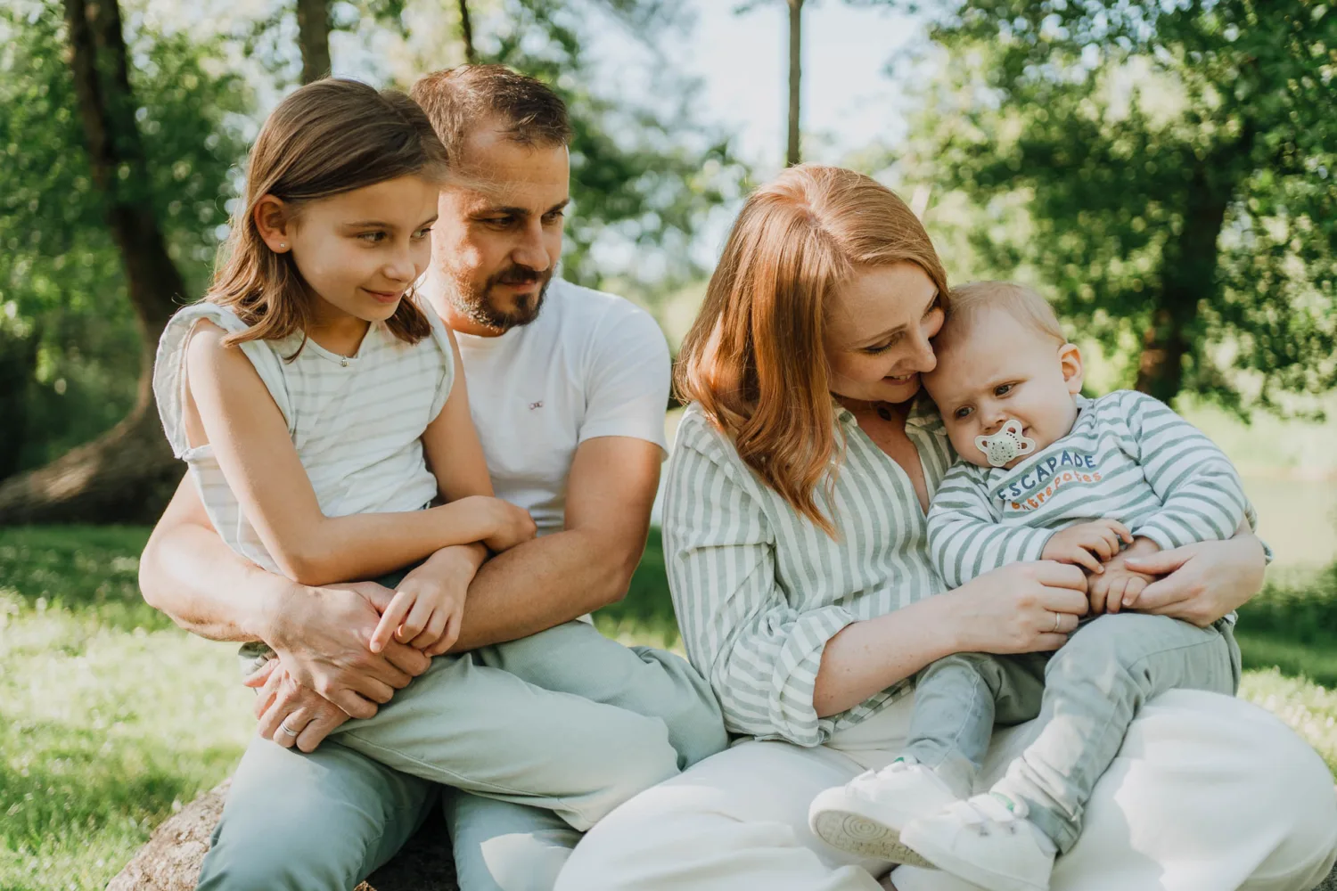 Une famille de quatre personnes est assise dehors par une journée ensoleillée dans les Landes. Le père tient l’aîné dans ses bras, tandis que la mère berce le bébé avec une tétine. Tous sont habillés de manière décontractée et entourés de verdure, capturant un parfait moment de photographe de famille.