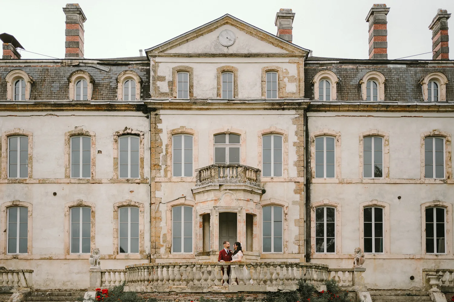 Un majestueux château français à trois étages, aux façades vieillies par le temps et doté d'un balcon central où se tient deux personnes, s'habille fièrement avec ses nombreuses fenêtres et cheminées en briques, offrant un cadre enchanteur idéal pour un photographe de mariage ou famille dans les pittoresques Landes.