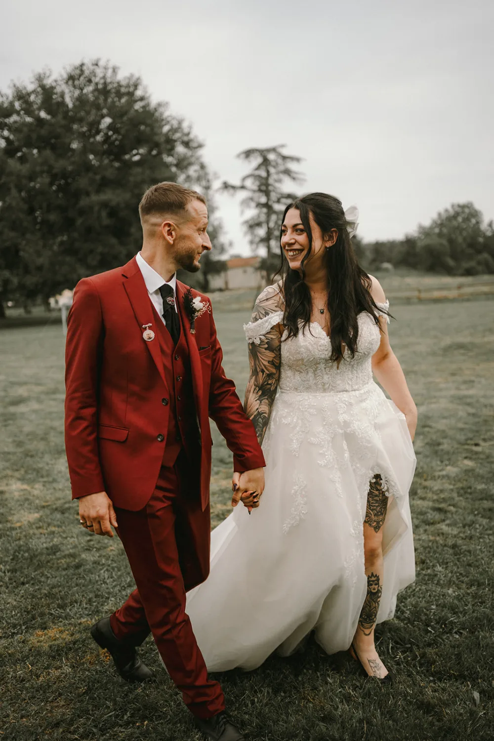 Un couple marchant main dans la main en plein air, l'homme vêtu d'un costume rouge et la femme portant une robe de mariée blanche, tous deux souriants comme immortalisés par un photographe de famille dans les Landes.