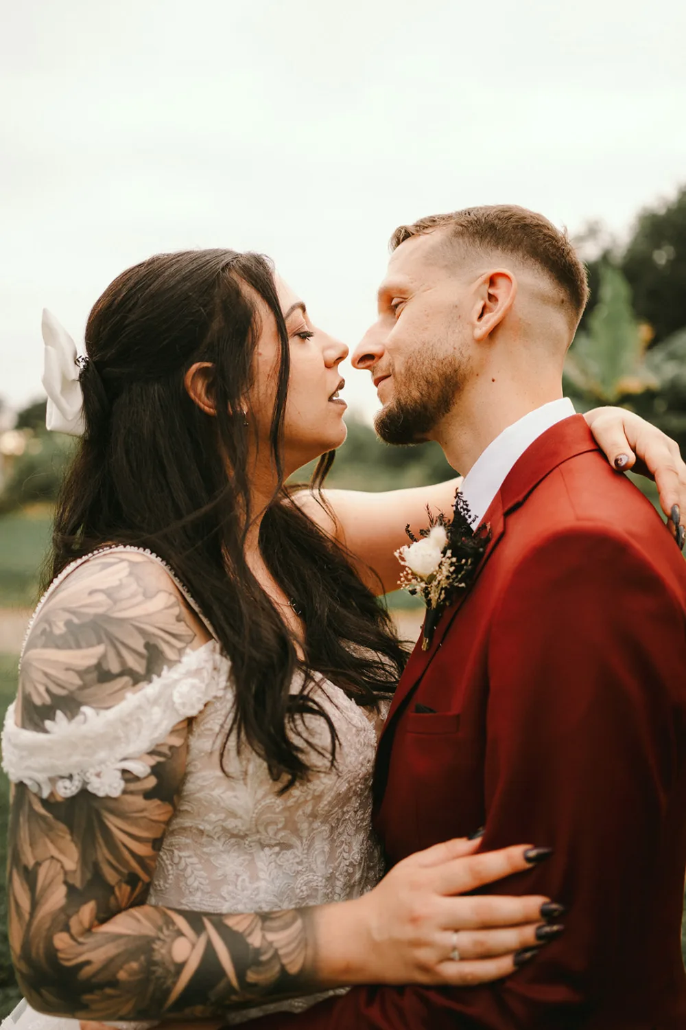 Un couple de mariés se tient tout près, se regardant tendrement dans les yeux, la mariée aux longs cheveux noirs arborant un tatouage floral sur le soutien-gorge et portant une robe en dentelle, tandis que le marié est vêtu d'un costume rouge élégant; cet instant magique est idéal pour un photographe de mariage ou de famille dans les Landes souhaitant immortaliser leur amour authentique.