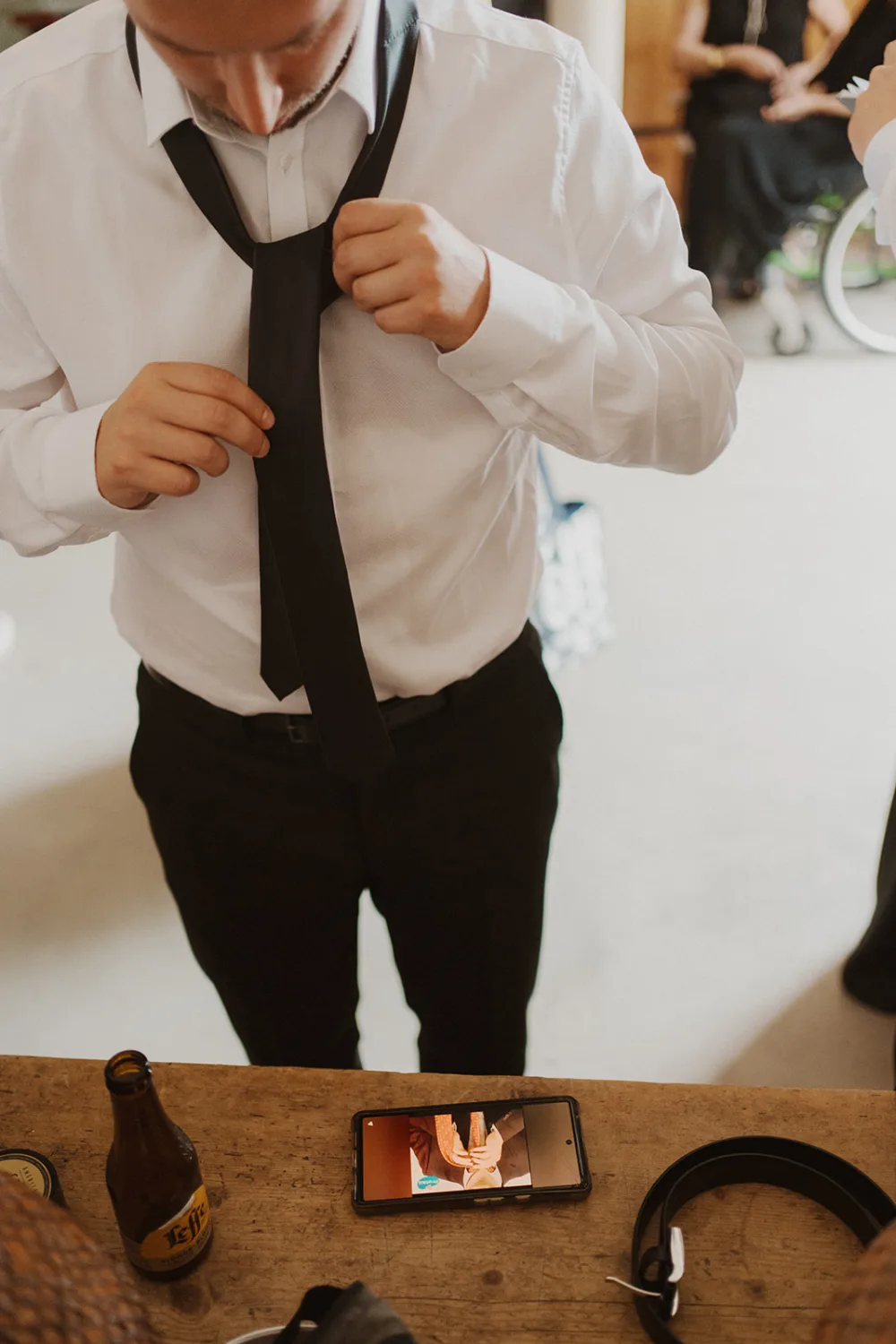 Un individu vêtu d'une chemise blanche et d'un pantalon noir ajuste soigneusement une cravate noire, tandis qu'en face de lui, sur la table, se trouve un smartphone diffusant un tutoriel vidéo pour nouer une cravate, une bouteille de bière et des écouteurs, évoquant les préparatifs méticuleux pour une séance photo chic avec un photographe de mariage ou de famille dans les Landes.