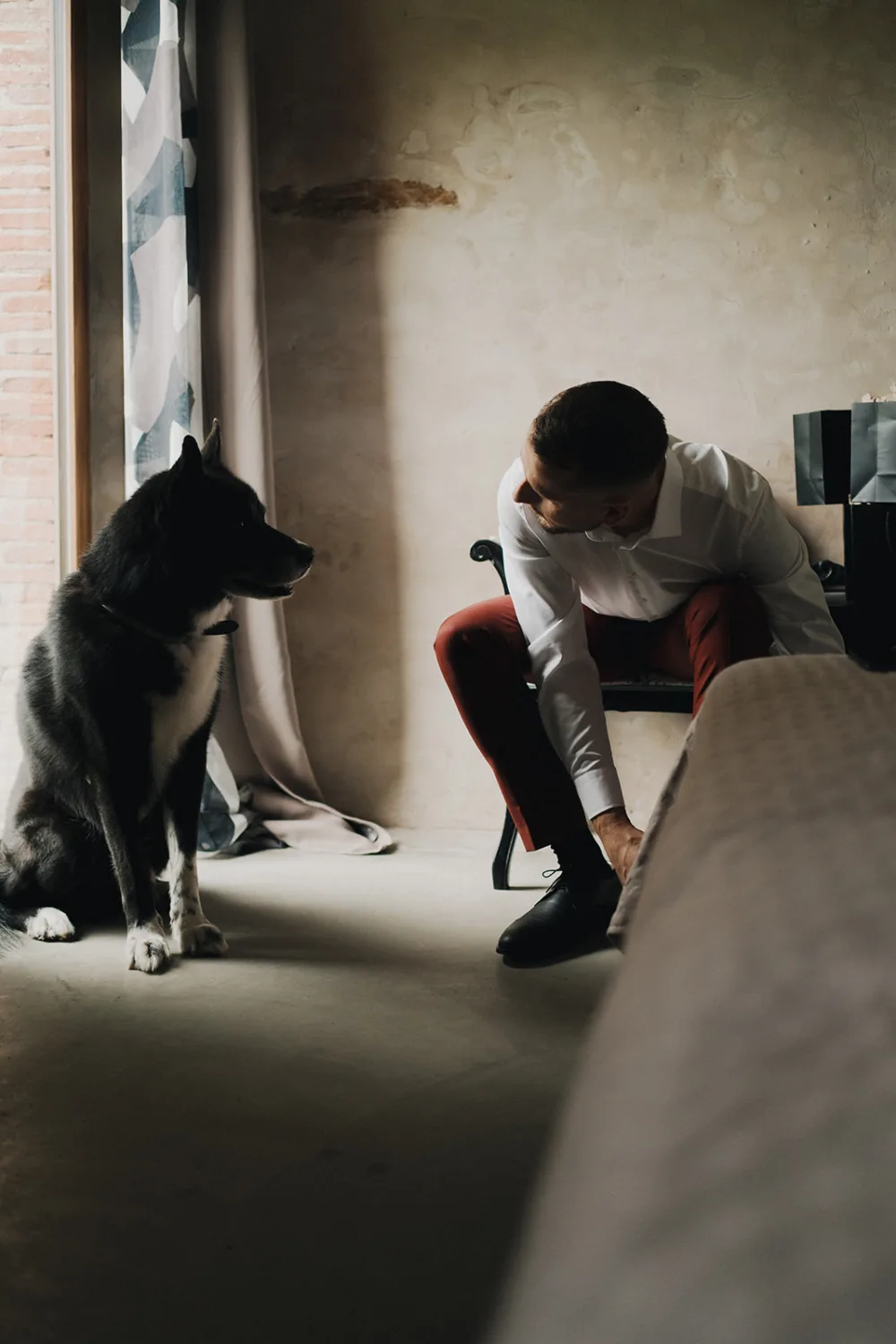 Dans une ambiance tamisée typique des séances de photographe famille Landes, un homme agenouillé ajuste ses lacets tandis qu'un chien noir et blanc, rappelant les nombreux compagnons à quatre pattes souvent capturés par les photographes de mariage et famille dans cette région pittoresque des Landes, observez attentivement la scène.