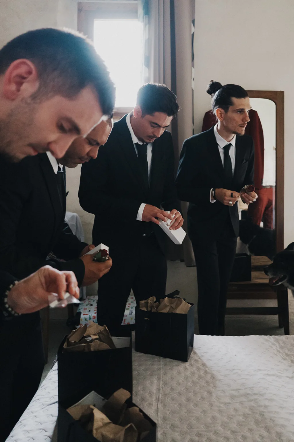 Lors d'un moment unique capturé par un *photographe de mariage et famille dans les Landes*, un groupe d'hommes élégamment vêtus en costumes se trouvent autour d'un lit, certains s'occupant avec des boîtes-cadeaux et divers objets ; la scène est sublimée par un chien assis près d'un homme agenouillé, tandis qu'à l'arrière-plan, un miroir et une fenêtre ajoutent une touche d'intimité à cette scène mémorable.