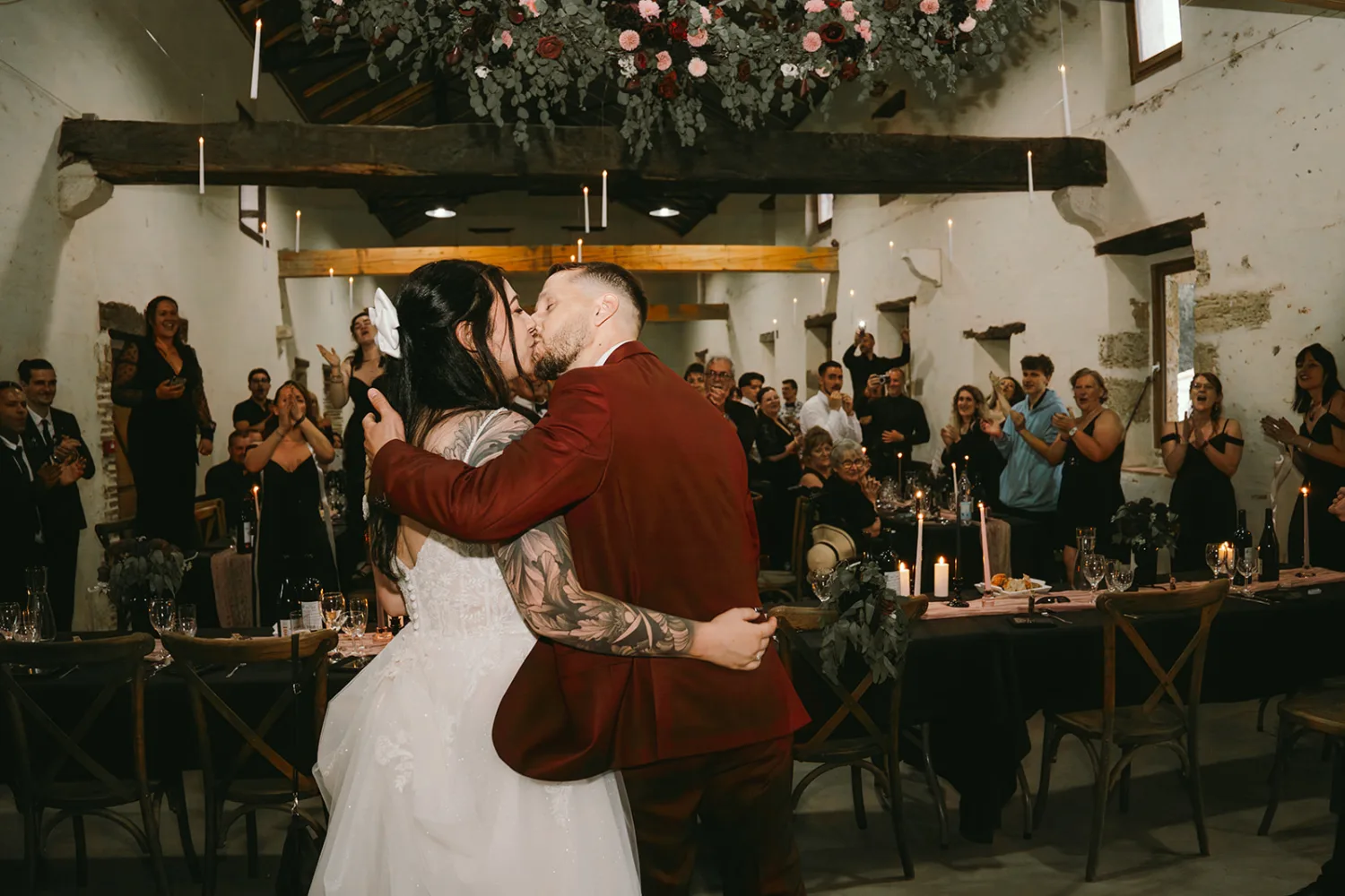 Lors de la cérémonie de mariage dans une salle rustique des Landes, un couple échange un baiser passionné entouré de leurs invités applaudissant et acclamant, ce moment mémorable étant capturé avec talent par le photographe famille Landes.