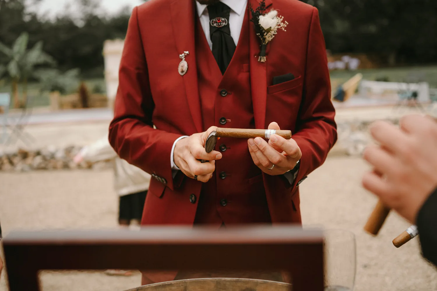 Lors d'un moment unique immortalisé par un photographe de mariage ou famille dans les Landes, une personne vêtue d'une élégante veste rouge et d'un gilet noir tient un cigare et un coupe-cigare, tout en recevant un autre cigare des mains. invisibles d'une tierce personne, créant ainsi une scène riche en détails et émotions.