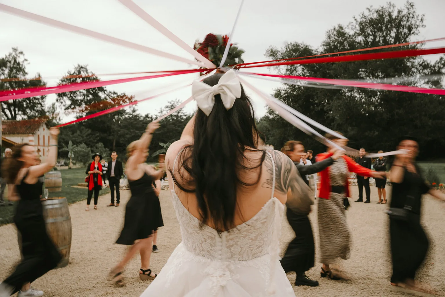Sur cette magnifique photo capturée par un photographe de mariage et famille dans les Landes, on voit une mariée ravissante ornée d'un grand nœud blanc sur la tête, tenant des rubans rouges et blancs, entourée d'invités joyeux qui dansent en plein air, créant une ambiance festive et mémorable.