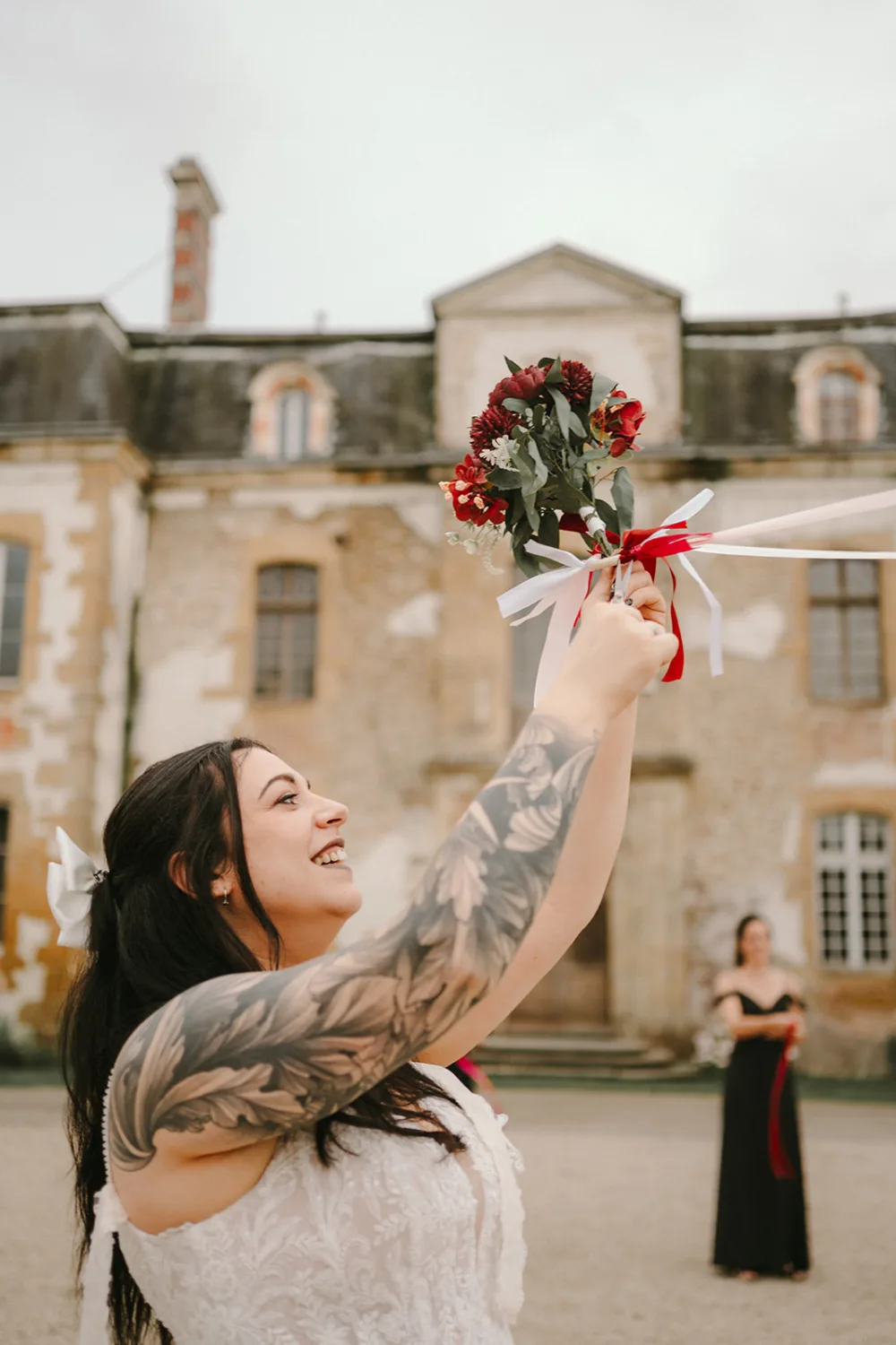 Une femme arborant des tatouages floraux et vêtue d'une robe blanche brandit un bouquet rouge et blanc, tandis qu'une autre personne se tient en arrière-plan devant un bâtiment historique, le tout capturé avec une grande finesse par un photographe de mariage et famille dans les Landes.