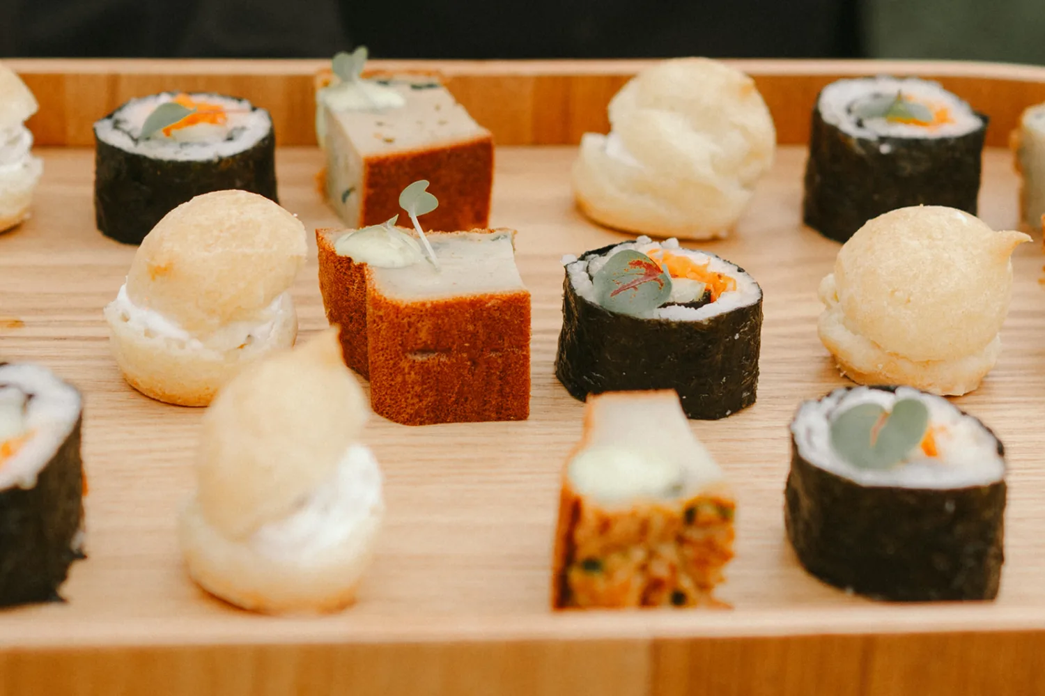 Un plateau en bois magnifiquement agence présente une variété d'amuse-bouches élégantes, comprenant des makis de sushi, des feuilletés dorés et des bouchées cubiques garnies de micro-pousses fraîches — une mise en scène idéale pour qu'un photographe famille Landes puisse immortaliser ces moments joyeux et délicats lors d'un événement ou mariage dans les Landes.