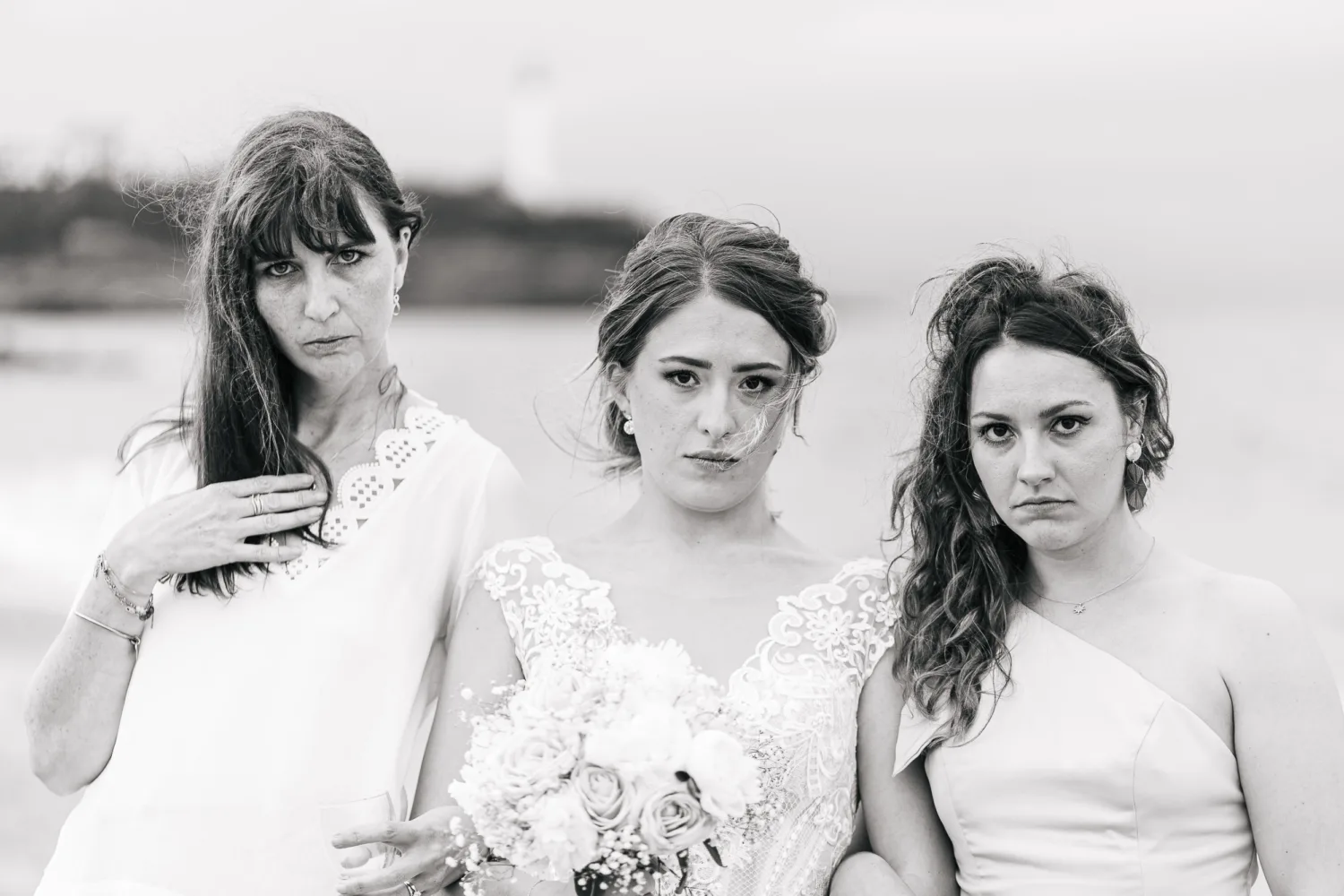 Sur une plage des Landes, trois femmes se respectent en rang avec des expressions sérieuses, la femme centrale portant une robe de mariée et tenant un bouquet tandis que les deux autres sont vêtues de manière décontractée et semi-formelle respectivement, capturant ainsi un moment parfait. et intemporel grâce à l'expertise d'un photographe mariage famille Landes au milieu d'un décor côtier serein.