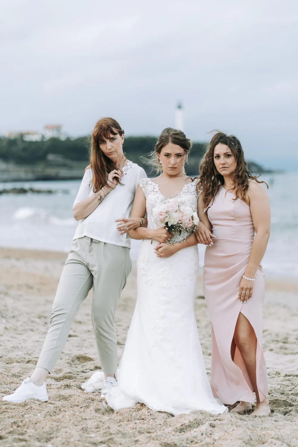 Découvrez une scène familiale émouvante où trois femmes rayonnent sur une plage des Landes, l'une élégamment vêtue d'une robe de mariée blanche, entourée à sa gauche par une femme en tenue décontractée et à sa droite par une autre en tenue de soirée chic , tandis que la mer scintillante et un phare pittoresque subliment ce moment parfait immortalisé par un photographe de mariage et famille dans les Landes.