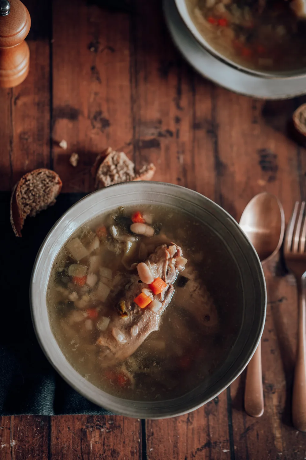 Une délicieuse soupe réconfortante avec une cuisse de poulet, des légumes en dés, des haricots et des verts, magnifiquement disposée sur une table en bois avec une cuillère, une fourchette et des morceaux de pain—évoquant les repas familiaux chaleureux pendant les vacances à Porto —parfaite pour un reportage photographique de mariage ou de moments en famille dans les Landes.